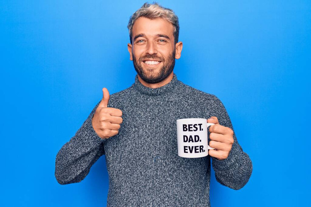 Handsome blond man drinking cup of coffee with best dad ever message over blue background smiling happy and positive, thumb up doing excellent and approval sign - Photo, Image