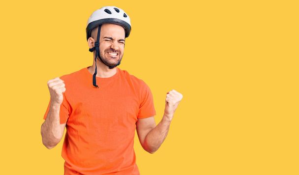 Giovane bell'uomo che indossa il casco da bici molto felice ed eccitato facendo il gesto del vincitore con le braccia alzate, sorridente e urlando per il successo. concetto di celebrazione.  - Foto, immagini