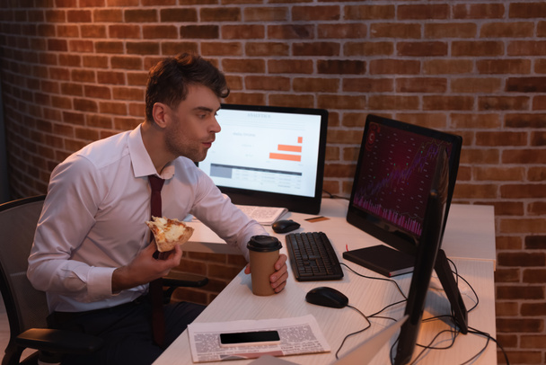 hombre de negocios comiendo pizza con café para llevar mientras revisa las existencias de computadoras en la oficina  - Foto, imagen