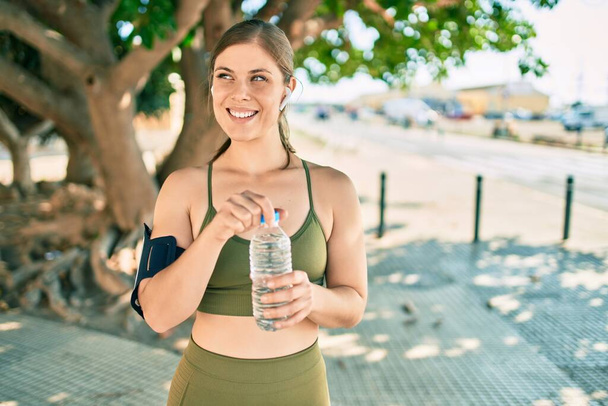 Young blonde sportswoman doing exercise drinking bottle of water at the city. - Photo, Image