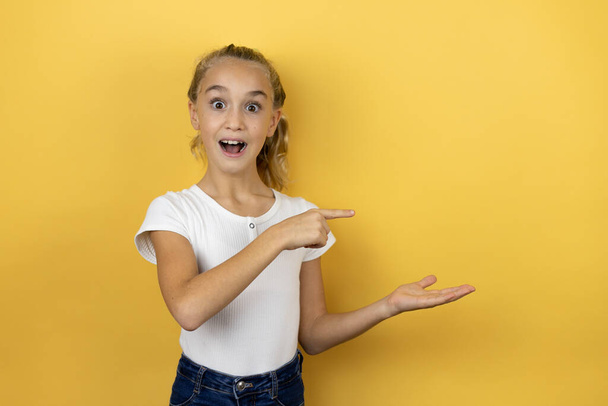 Young beautiful child girl standing over isolated yellow background surprised, showing and pointing something that is on her hand - Fotoğraf, Görsel