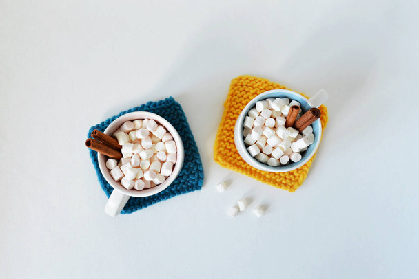 Mustard yellow and dark blue handmade knitted coasters, cups with marshmallows and cinnamon over white - 写真・画像