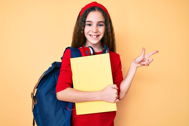 Linda niña hispana usando mochila estudiantil y auriculares sosteniendo libro sonriendo feliz señalando con la mano y el dedo al lado  - Foto, imagen