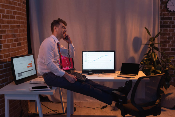 Mosolygó üzletember beszél telefonon közelében eszközök és számítógépek este - Fotó, kép