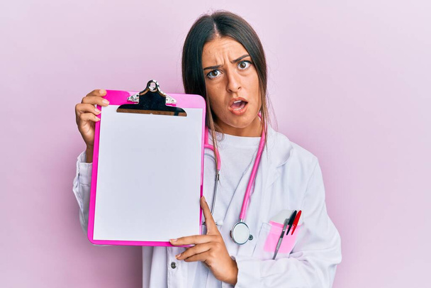 Krásná hispánská žena v lékařském stetoskopu drží podložku v šokovém obličeji, vypadá skepticky a sarkasticky, překvapen s otevřenými ústy  - Fotografie, Obrázek