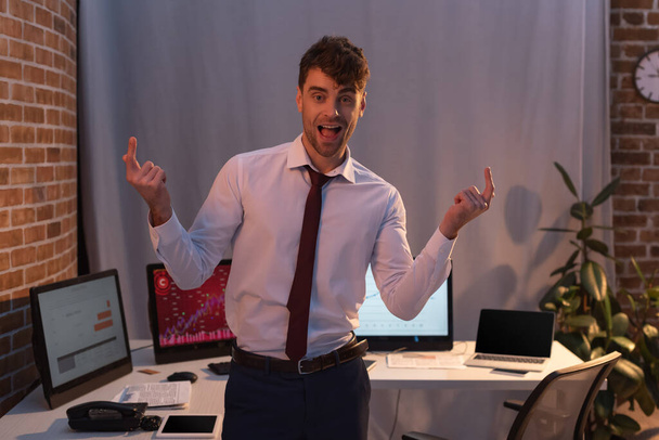 Fröhlicher Geschäftsmann zeigt mit Fingern in der Nähe von Computern auf verschwommenem Hintergrund  - Foto, Bild