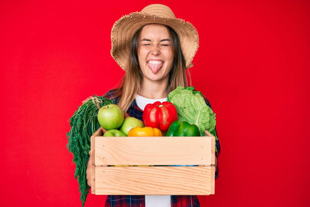Beautiful caucasian woman wearing farmer hat holding vegetables sticking tongue out happy with funny expression.  - Photo, Image
