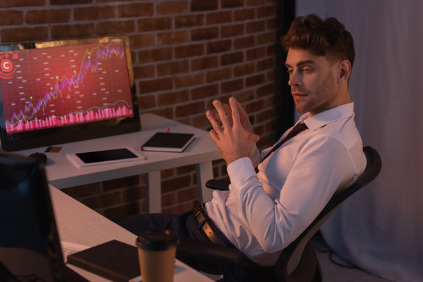 businessman sitting near computers with charts and coffee to go on blurred foreground in evening  - Photo, Image