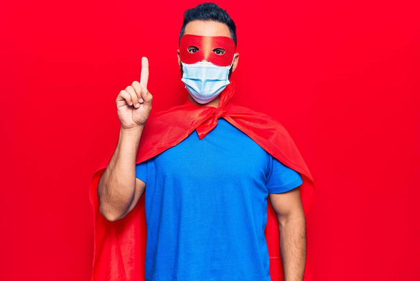 Joven hombre hispano con disfraz de superhéroe y máscara médica sonriendo con una idea o pregunta apuntando con el dedo con cara feliz, número uno  - Foto, Imagen