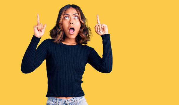 Young beautiful mixed race woman wearing casual clothes amazed and surprised looking up and pointing with fingers and raised arms.  - Photo, Image
