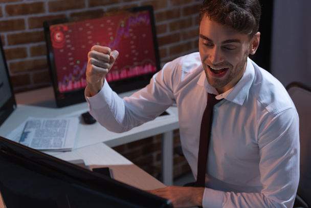 businessman showing yeah gesture near computer on blurred foreground in evening  - Photo, Image