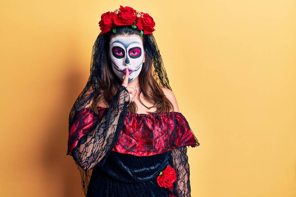Young woman wearing day of the dead costume over yellow asking to be quiet with finger on lips. silence and secret concept.  - 写真・画像