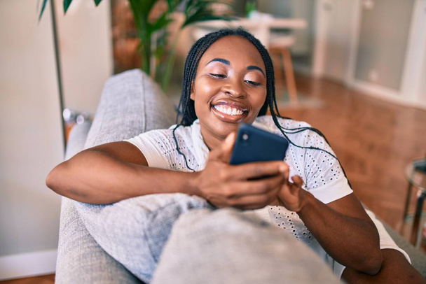 Молода афроамериканська жінка, яка посміхається і радіє, сидячи вдома за допомогою смартфона. - Фото, зображення