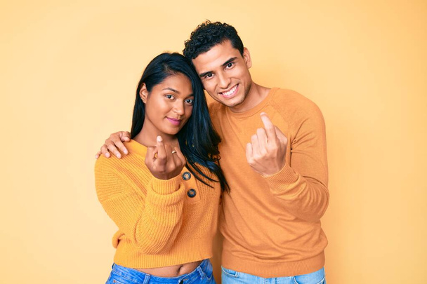 Gyönyörű latin fiatal pár viselnek alkalmi ruhák együtt integetve gyere ide gesztus kézzel hívogató üdvözlő boldog és mosolygós  - Fotó, kép