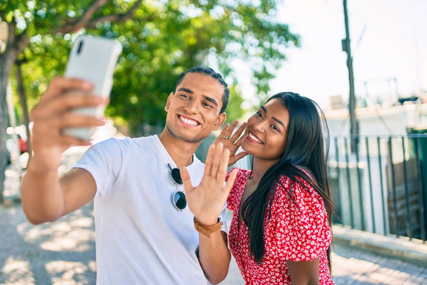 若いラテンカップルは、街の通りでビデオ通話のためのスマートフォンを使用して幸せな笑顔. - 写真・画像