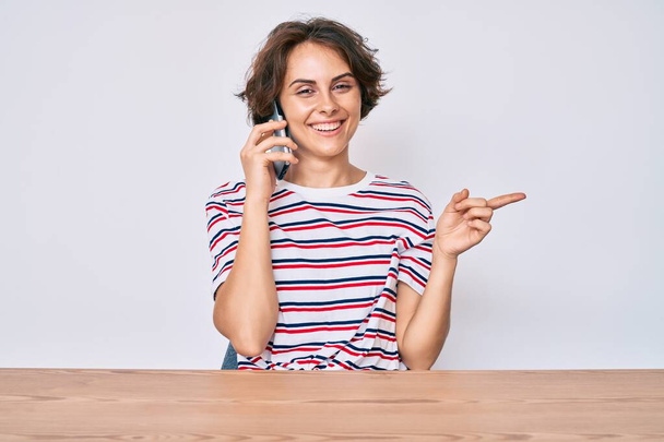 Молода іспаномовна жінка розмовляє на смартфоні, сидячи на столі, усміхаючись, щасливо вказуючи рукою і пальцем на бік
  - Фото, зображення