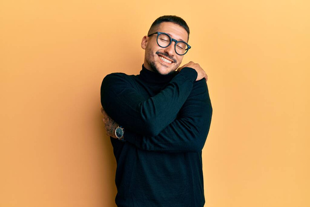 Pohledný muž s tetováním v roláku svetru a brýlích objímající se šťastný a pozitivní, usmívající se sebevědomě. self love a self care  - Fotografie, Obrázek
