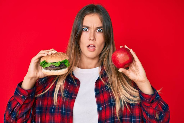Όμορφη καυκάσια γυναίκα σύγκριση burger και υγιή κόκκινο μήλο στο πρόσωπο σοκ, αναζητούν δύσπιστοι και σαρκαστικός, έκπληκτος με ανοιχτό στόμα  - Φωτογραφία, εικόνα