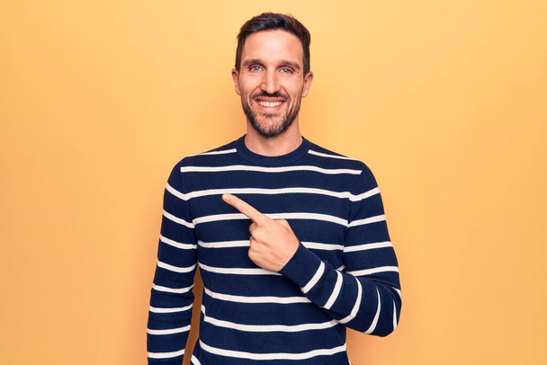 Young handsome man wearing casual striped sweater standing over isolated yellow background smiling cheerful pointing with hand and finger up to the side - Photo, Image