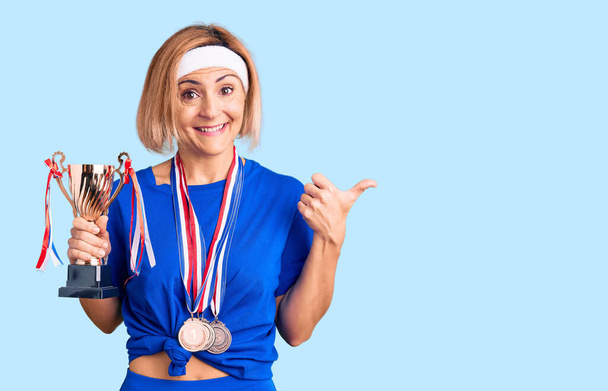 Joven mujer rubia sosteniendo trofeo campeón llevando medallas apuntando el pulgar hacia el lado sonriendo feliz con la boca abierta  - Foto, imagen