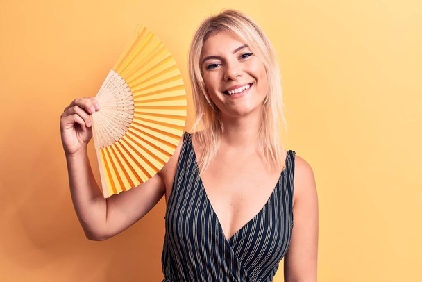 Junge schöne blonde Frau mit Handventilator, der über isoliertem gelben Hintergrund steht, sieht positiv und glücklich stehend aus und lächelt mit einem selbstbewussten Lächeln, das Zähne zeigt - Foto, Bild
