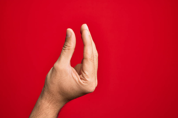 Ręka białego młodzieńca pokazuje palce nad odizolowanym czerwonym tłem zbierając i biorąc niewidzialną rzecz, trzymając obiekt z palcami pokazującymi przestrzeń - Zdjęcie, obraz