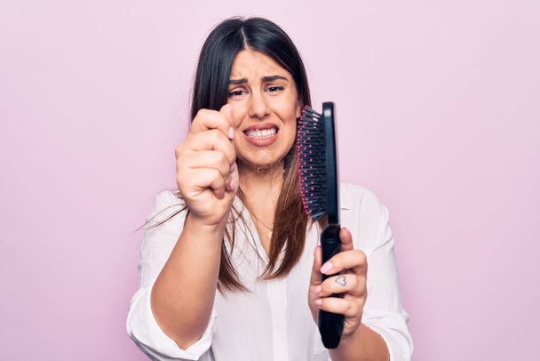 Junge schöne Frau agil und besorgt um Kapillarprobleme. Haarbürste mit wirren Haaren vor isoliertem rosa Hintergrund halten - Foto, Bild