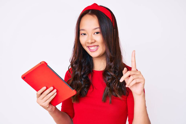 Молодая красивая китайская девушка держит тачпад удивлен идеей или вопрос указывая пальцем со счастливым лицом, номер один  - Фото, изображение