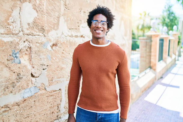 Νεαρός όμορφος Αφροαμερικάνος που φοράει καθημερινά ρούχα και γυαλιά χαμογελώντας χαρούμενος. Στέκεται με χαμόγελο στο πρόσωπο κοιτάζοντας την κάμερα περπατώντας στο δρόμο της πόλης. - Φωτογραφία, εικόνα