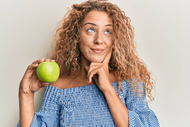 Kaunis valkoihoinen teini-ikäinen tyttö, jolla on vihreä omena vakava kasvot ajattelevat kysymystä käsin leualla, huomaavainen hämmentävästä ideasta.  - Valokuva, kuva