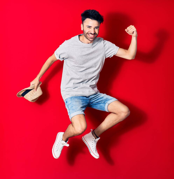 Young handsome hispanic man wearing casual clothes smiling happy. Jumping with smile on face holding summer hat celebrating with fist up over isolated red background. - Foto, Bild