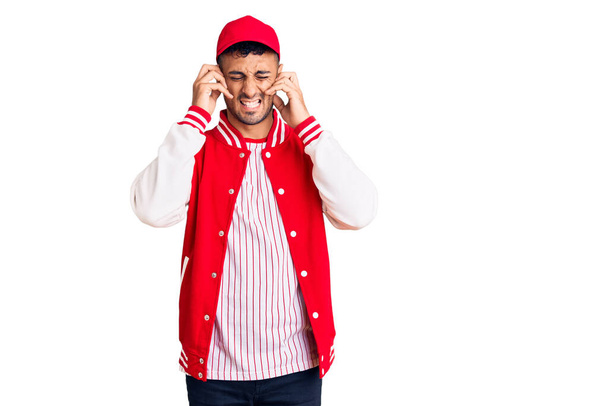 Young hispanic man wearing baseball uniform covering ears with fingers with annoyed expression for the noise of loud music. deaf concept.  - 写真・画像