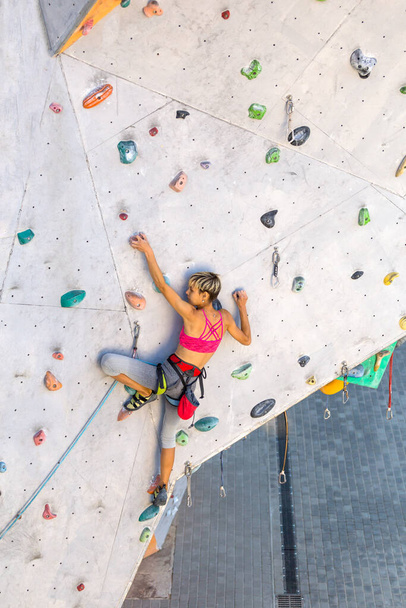 Een vrouw klimt over een klimwand, een klimmer traint op kunstmatig terrein, rotsklimmen in de stad, een sterk meisje, sport in de stad, veiligheid in extreme sporten. - Foto, afbeelding