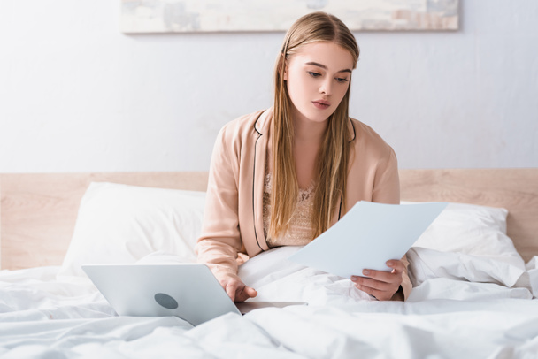 young freelancer in silk robe looking at documents near laptop on bed - Foto, Bild