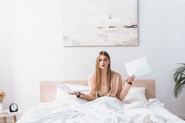 confused young freelancer in silk robe holding laptop and documents in bedroom - Фото, изображение