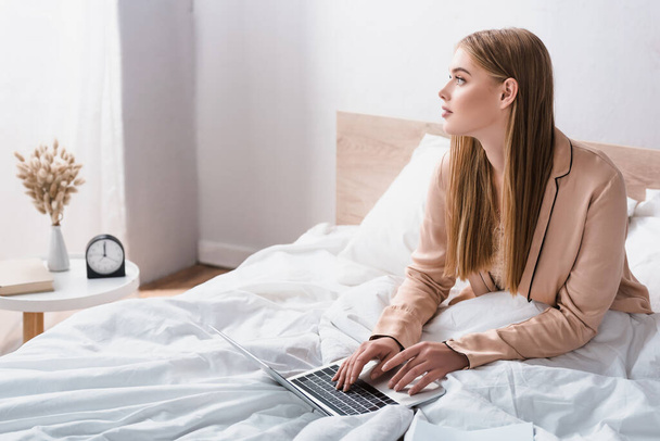 young woman in silk robe using laptop and looking away on bed - Foto, Imagen