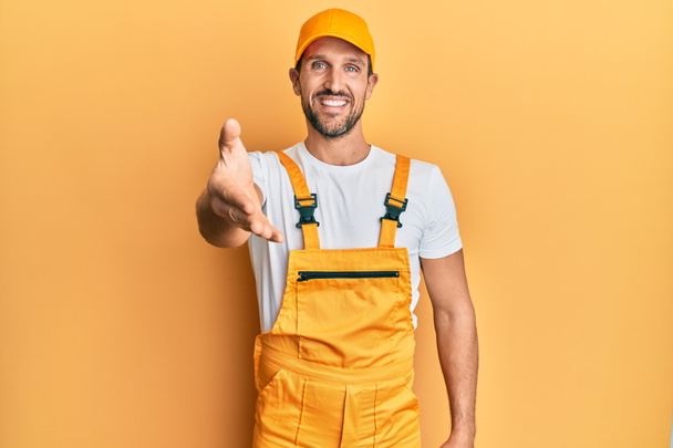 Young handsome man wearing handyman uniform over yellow background smiling friendly offering handshake as greeting and welcoming. successful business.  - Photo, Image
