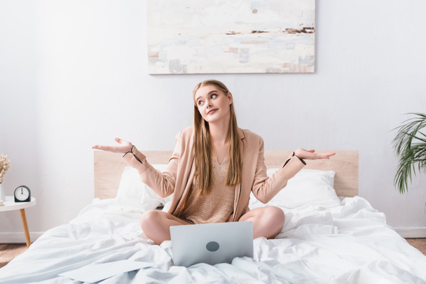 confused young woman in satin robe looking away near laptop on bed - Photo, Image