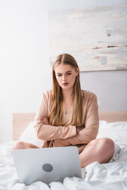 young woman sitting with crossed arms and looking at laptop on bed - Foto, Imagen