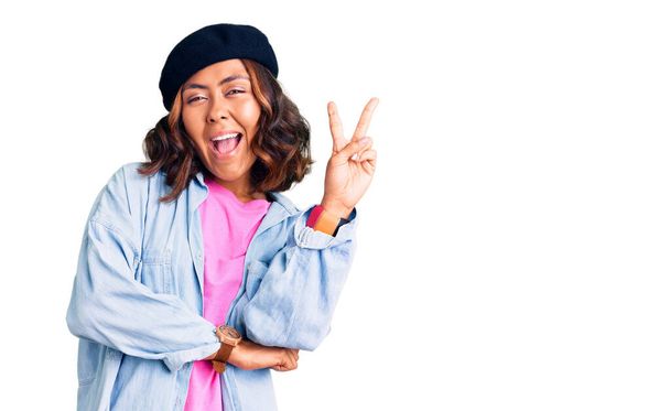 Young beautiful mixed race woman wearing french look with beret smiling with happy face winking at the camera doing victory sign. number two.  - Photo, Image