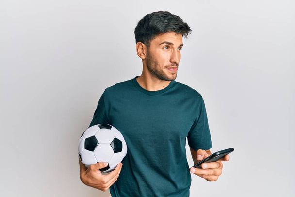 Jonge knappe man met voetbal bal kijken naar smartphone bet app glimlachen naar de zijkant en staren weg denken.  - Foto, afbeelding