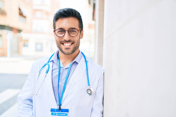 Young handsome hispanic doctor wearing uniform and stethoscope smiling happy. Standing with smile on face leaning on the wall at town street. - Foto, Bild