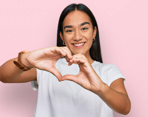 Mujer asiática joven vistiendo camiseta blanca casual sonriendo en el amor haciendo forma de símbolo del corazón con las manos. concepto romántico.  - Foto, imagen