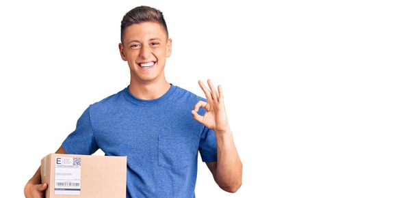 Homem hispânico bonito jovem segurando pacote de entrega fazendo sinal ok com os dedos, sorrindo simpática gesticulando excelente símbolo  - Foto, Imagem