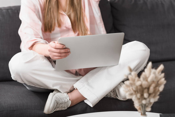 cropped view of young freelancer sitting with crossed legs on couch and using laptop - Foto, Bild