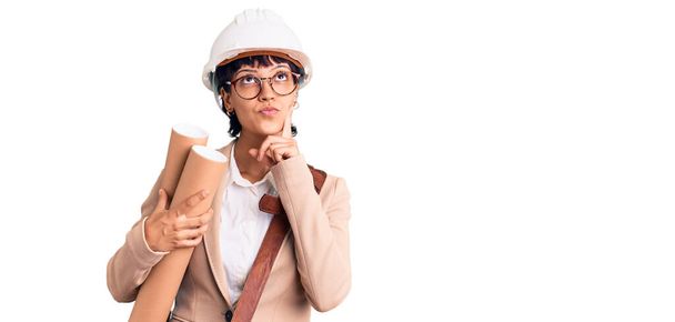 Young brunette woman with short hair wearing safety helmet holding blueprints serious face thinking about question with hand on chin, thoughtful about confusing idea  - Foto, Imagem