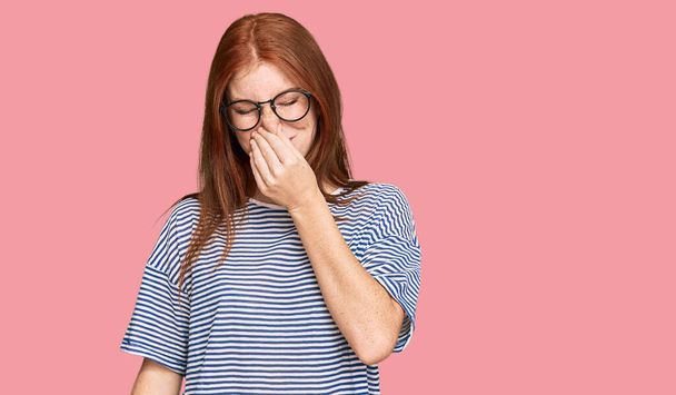 Young read head woman wearing casual clothes and glasses smelling something stinky and disgusting, intolerable smell, holding breath with fingers on nose. bad smell  - Foto, Imagen