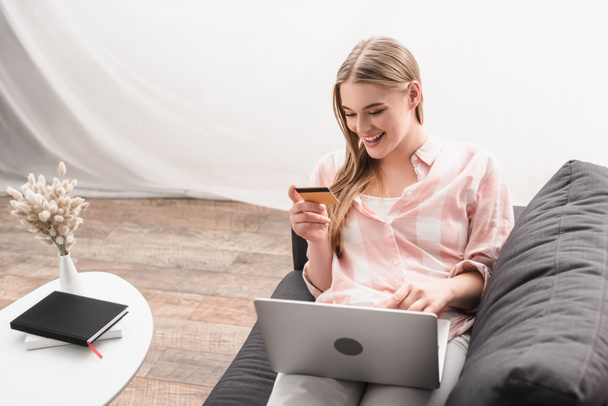 щаслива молода жінка тримає кредитну картку біля ноутбука, сидячи на дивані
 - Фото, зображення