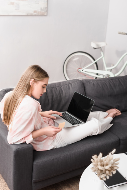 młoda kobieta trzyma kartę kredytową w pobliżu laptopa z pustym ekranem podczas e-zakupów w domu - Zdjęcie, obraz
