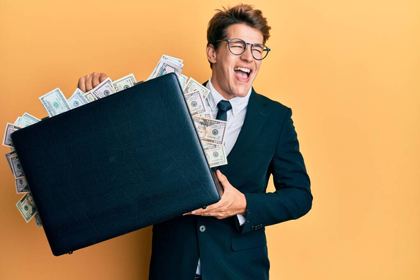 Handsome caucasian business man holding briefcase full of dollars celebrating crazy and amazed for success with open eyes screaming excited.  - Photo, Image